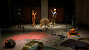 Two artists hold up wires, standing either side of a wooden crib-like structure on the floor - the photo captures a moment in Rita Evans' Tuning in a Vacuum piece
