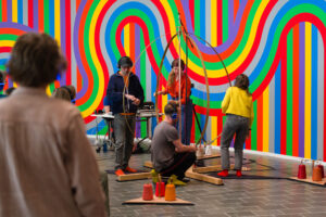 A group of performers work with large strips of copper in the shape of a large egg against a multicoloured background - the photo captures a moment in Rita Evans' Stringing the Matrix piece