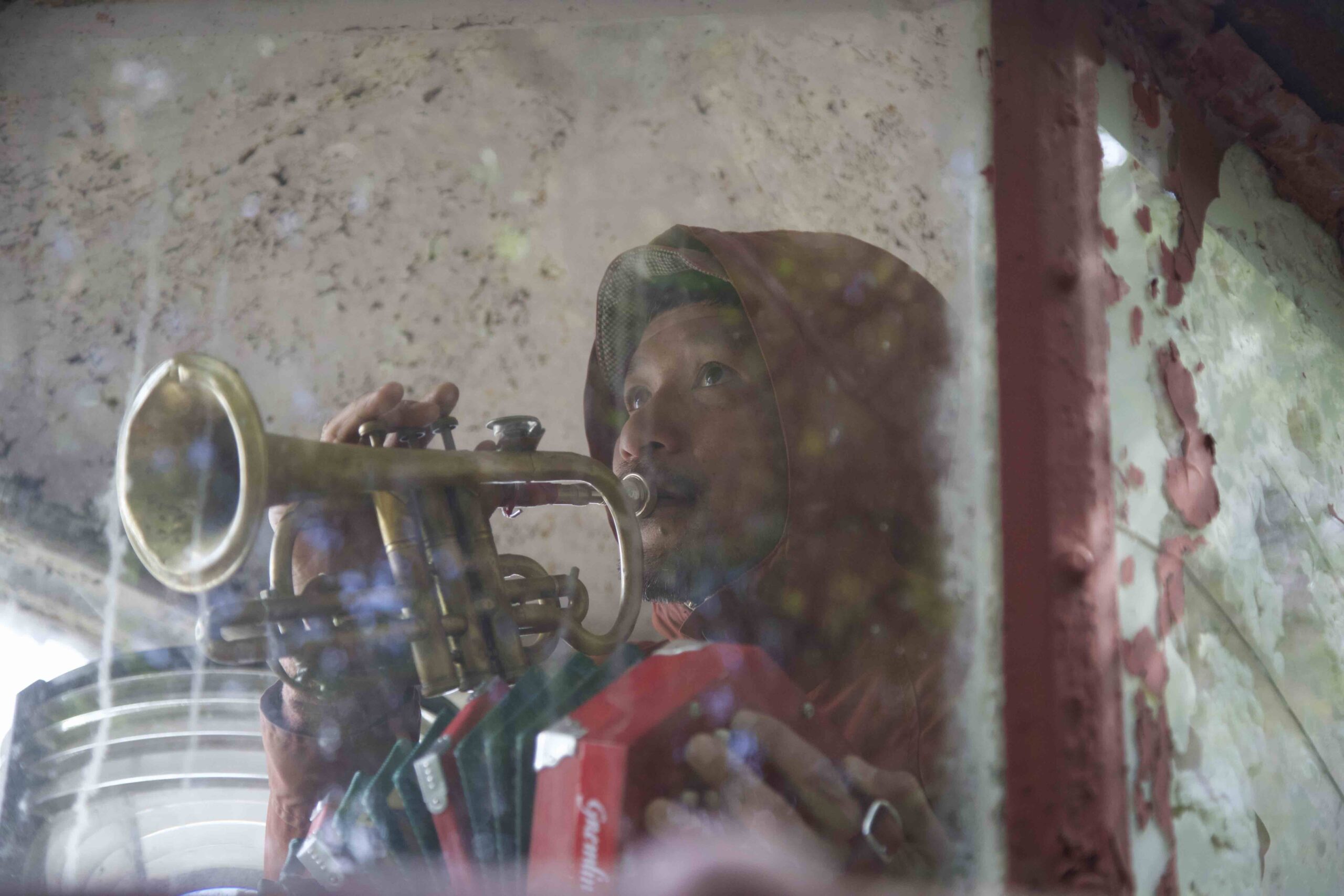 Ichi playing trumpet against wall with peeling red paint.